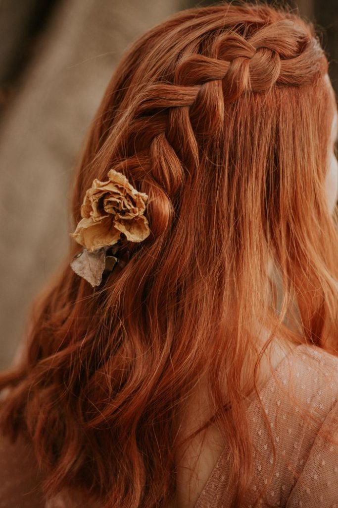 Red-haired woman with a braided hairstyle adorned with a dried flower, captured in a soft, warm tone.
